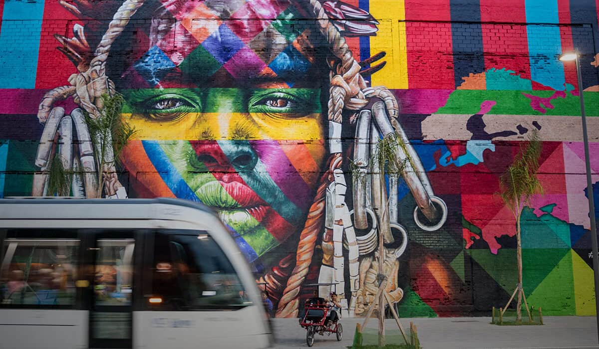 A colorful mural of a persons face covers a large wall. A white bus drives by, and there are planted trees in front of the mural.