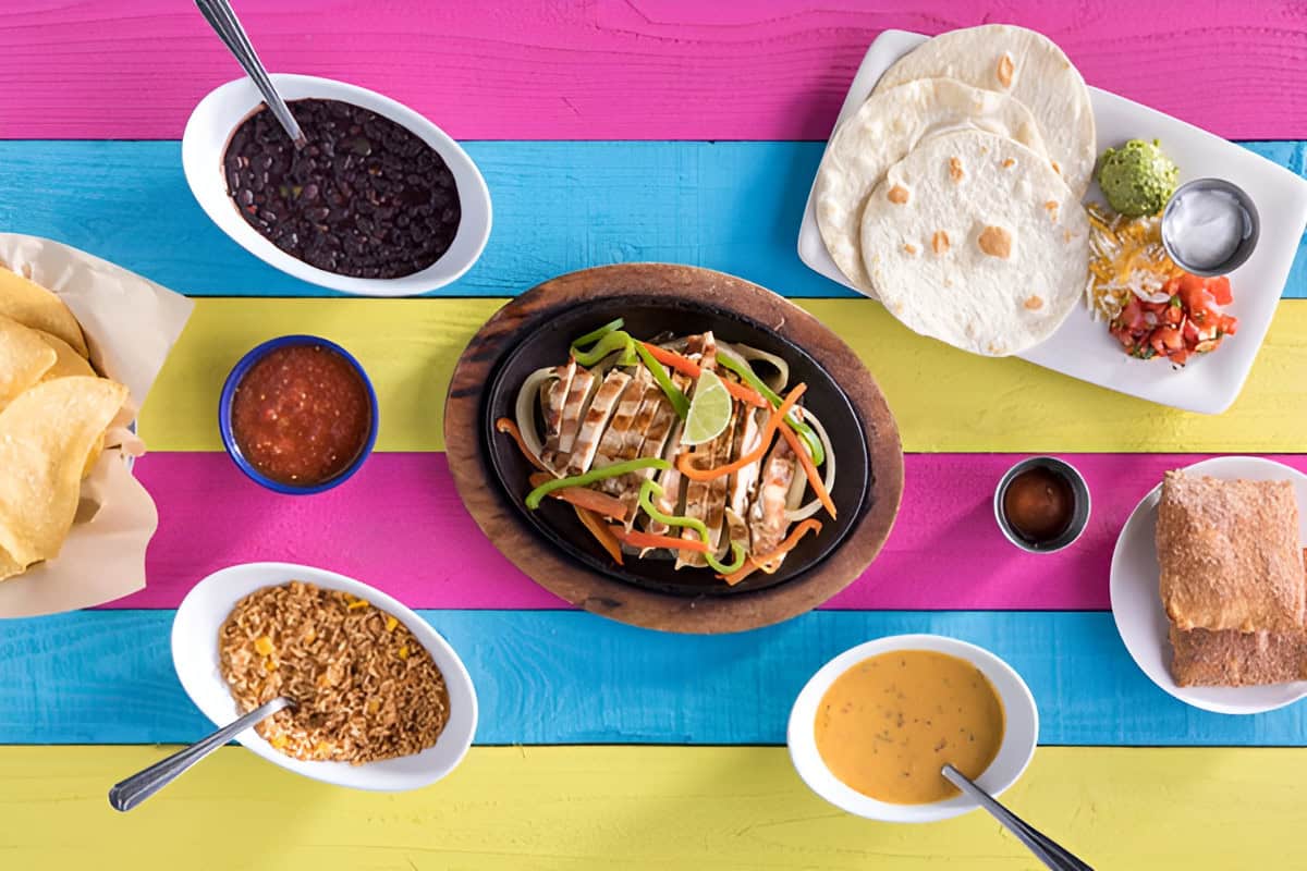 A colorful table setting with grilled chicken fajitas, tortillas, assorted salsas, beans, rice, queso, and tortilla chips.