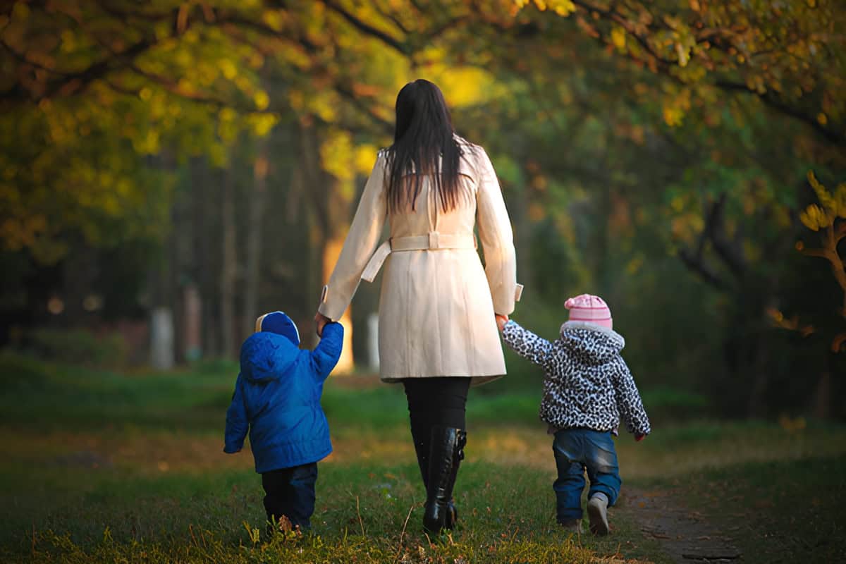 A woman in a white coat walks hand in hand with two children in colorful jackets through a park with autumn foliage.