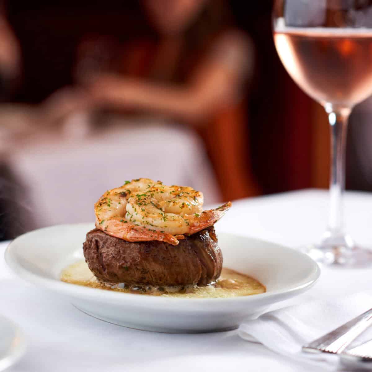 A plate of steak topped with shrimp is elegantly presented on a table, reminiscent of a Ruth’s Chris signature dish, with a glass of rosé in the background.