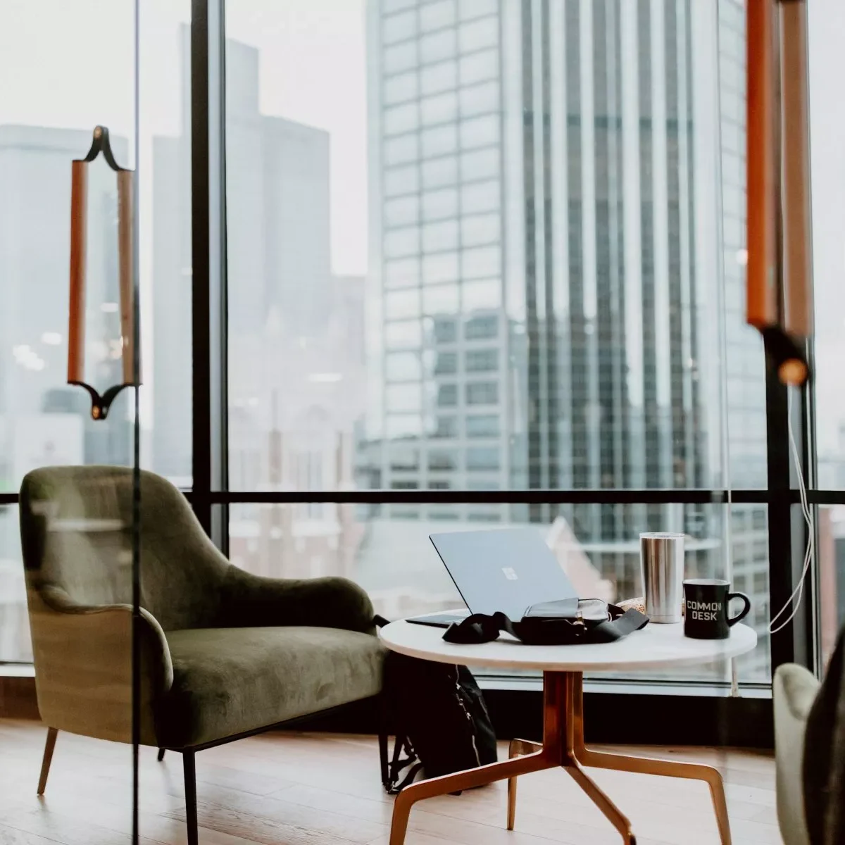 Modern office interior with a green armchair, round table featuring a laptop, headphones, and a mug. Large windows offer a view of tall city buildings.