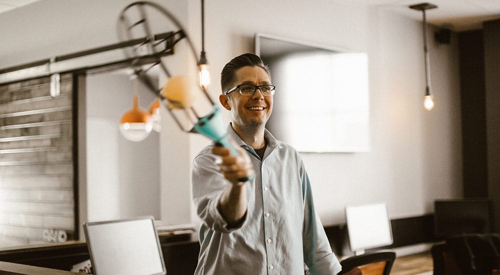 A man in a button-up shirt playfully holds out a device in an office with multiple computer monitors.