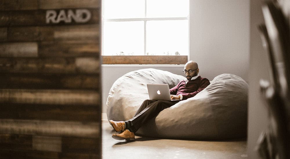 A person sitting on a large bean bag in a room, working on a laptop.