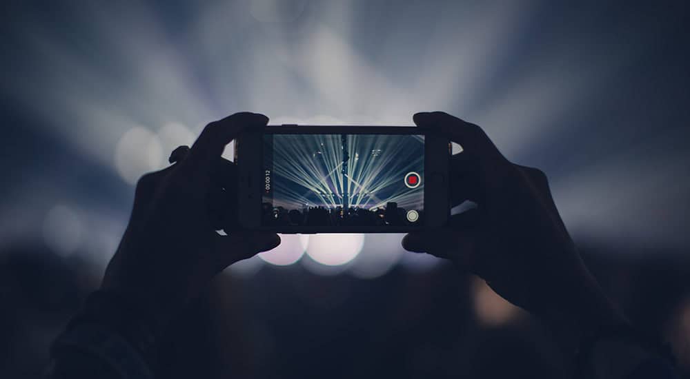 Person holding a smartphone recording a concert, with stage lights and a crowd visible on the screen.