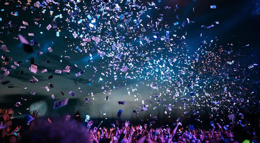 A crowd of people with raised hands at a concert under a night sky, with colorful confetti falling from above.