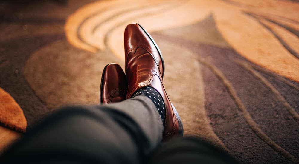Close-up of a persons feet wearing brown dress shoes and dotted socks, resting on a patterned carpet.