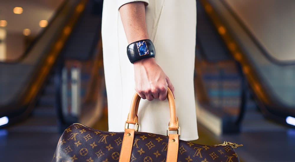 Person holding a brown designer handbag with tan handles, wearing a black smartwatch, standing in front of escalators.