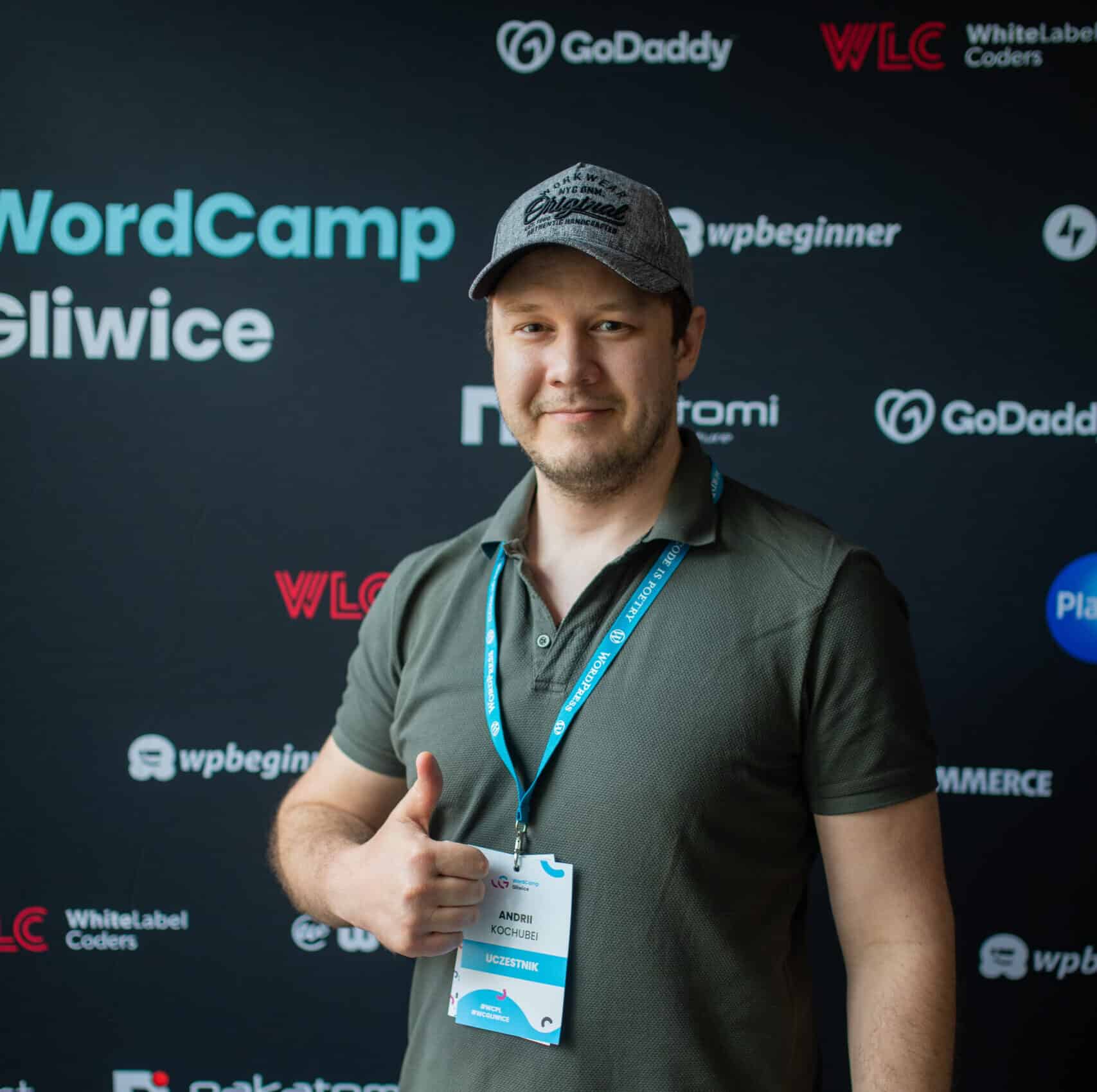 A person wearing a gray cap and green shirt gives a thumbs-up in front of a WordCamp Gliwice backdrop with various tech logos.