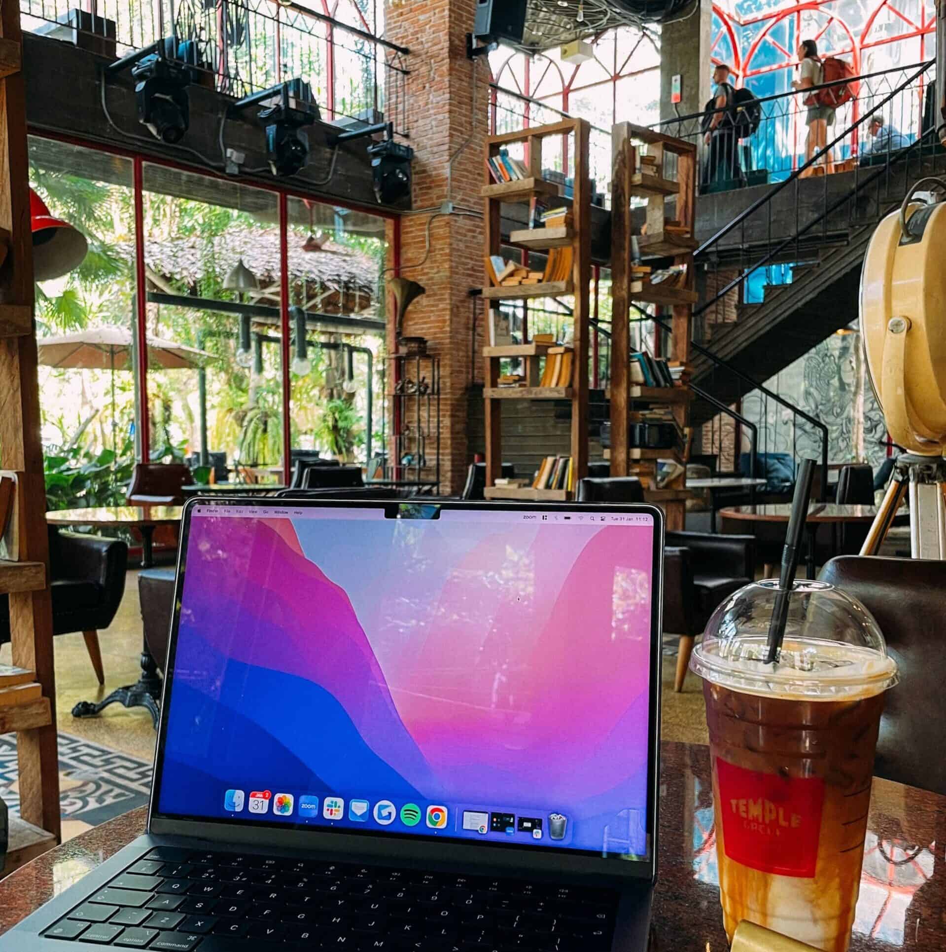 A laptop on a table in a cozy cafe with bookshelves and a staircase. Iced tea and a number stand are also on the table.