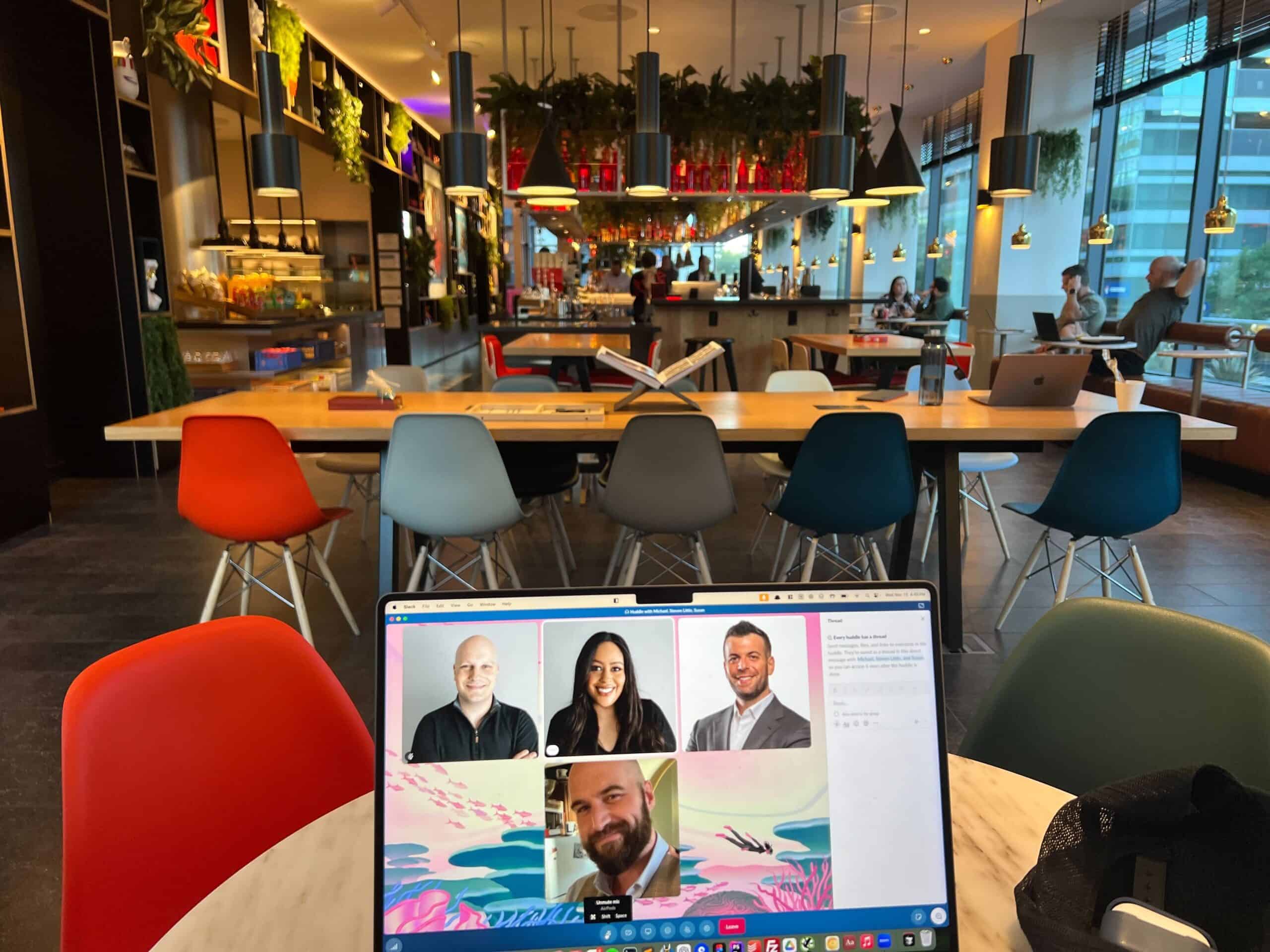 A laptop on a table shows a video call with four people. The setting is a modern café with colorful chairs, hanging lights, and plants.