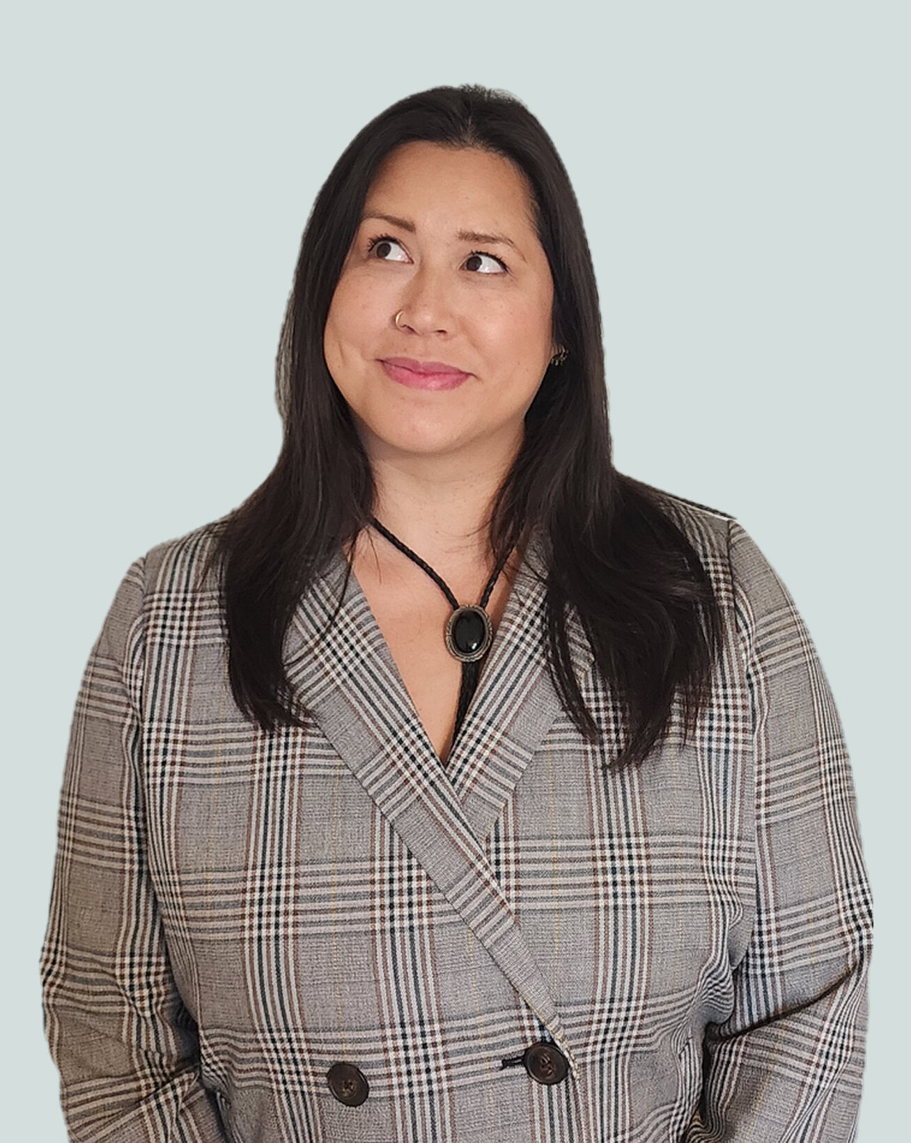Woman with long dark hair wearing a gray plaid blazer and a black bolo tie with an oval pendant, looking slightly upward and to the side with a subtle smile.