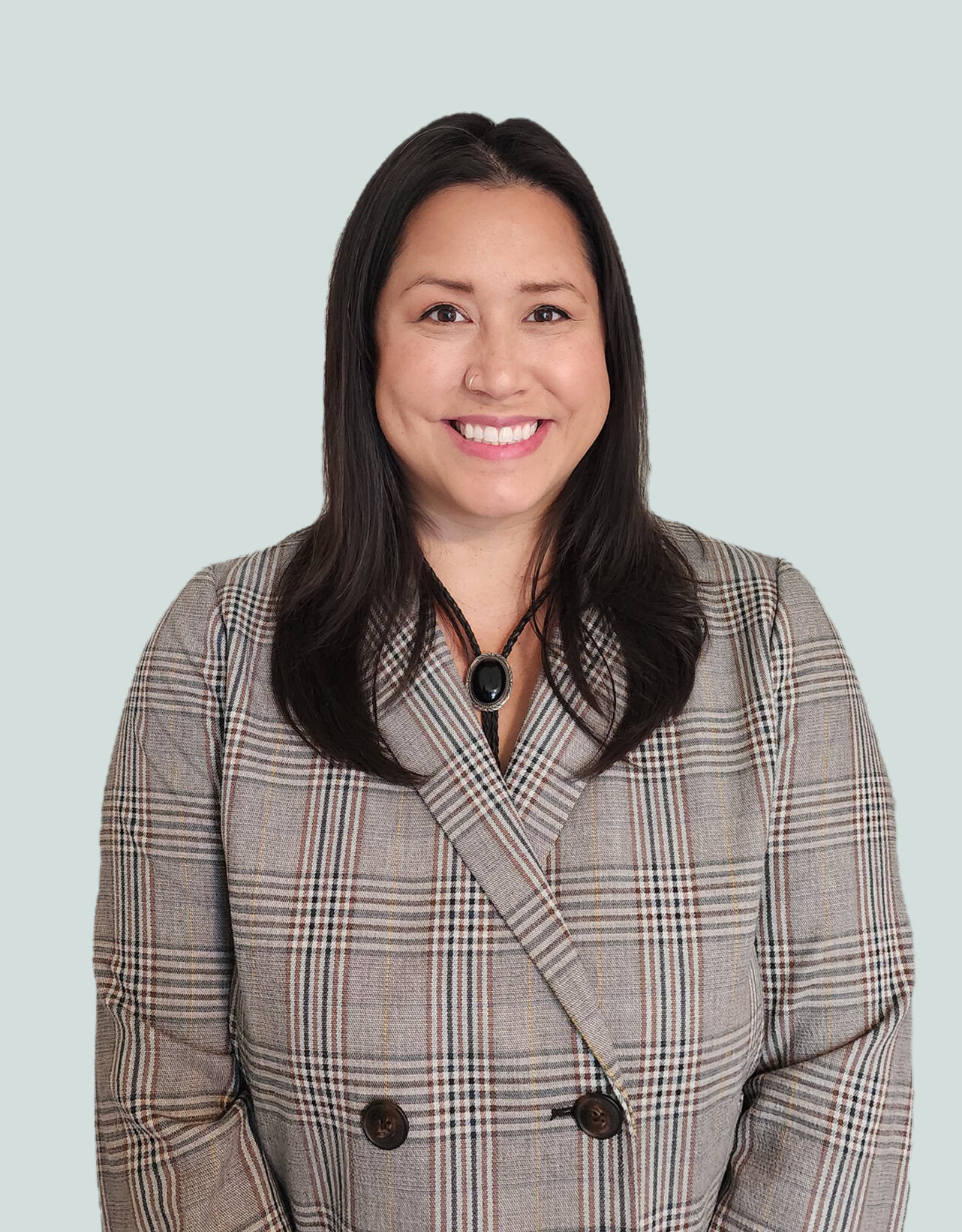 Woman with long dark hair wearing a gray plaid double-breasted blazer and a black bolo tie with a large oval stone, smiling against a light gray background.