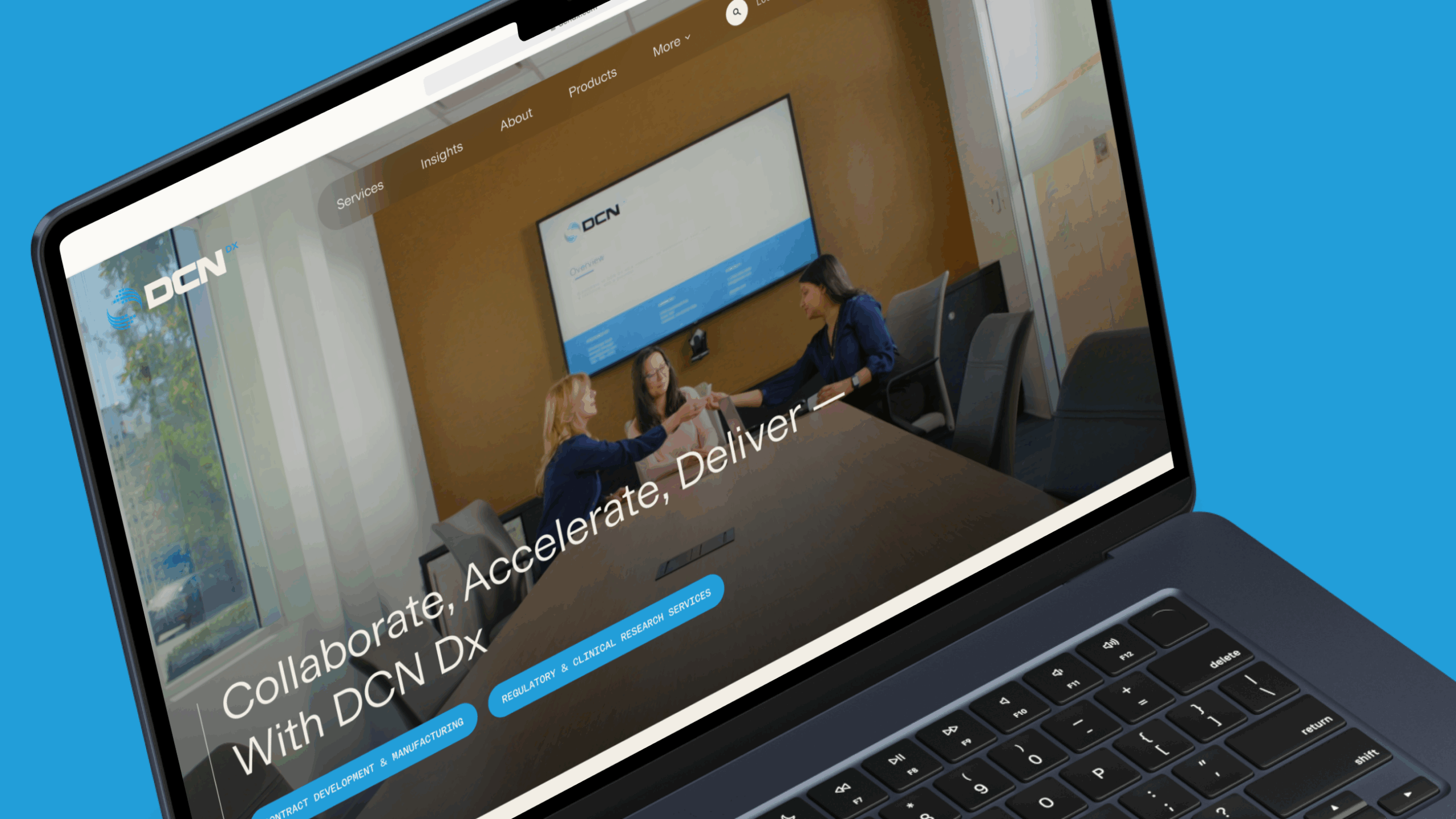 A laptop screen displays a website featuring three women in a conference room, sitting around a table and exchanging a document or item. The background wall is brown with a large screen showing the DCN logo and some text. The website header includes navigation links such as Services, Insights, About, Products, and More. The main text on the screen reads 