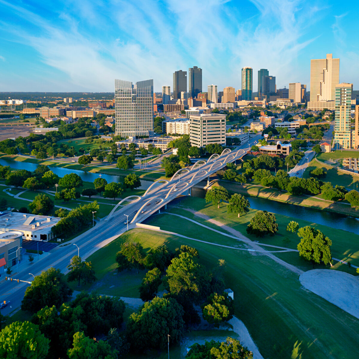 Aerial view of a modern city skyline with a distinctive arched bridge crossing a river. The scene includes numerous green trees and grassy areas surrounding the river and bridge, with various mid-rise and high-rise buildings in the background under a bright blue sky with wispy clouds.