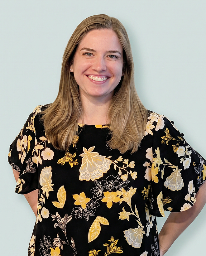 Woman with straight, shoulder-length blonde hair wearing a black blouse with yellow and white floral patterns, smiling against a light blue background.
