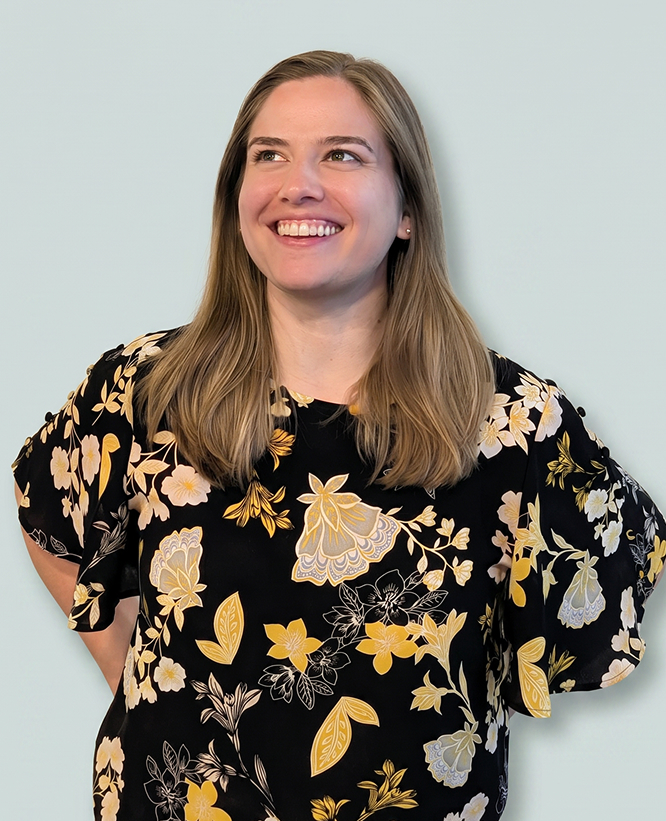 A woman with straight, shoulder-length blonde hair is smiling and looking slightly upward. She is wearing a black blouse with a floral pattern in shades of yellow, white, and beige. The background is a plain light gray color.